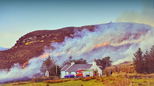 A fire appliance parked next to a small white house in the countryside, which is surrounded by hills on fire