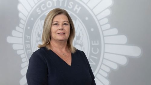 Liz Barnes in a black top standing in front of a grey SFRS crest