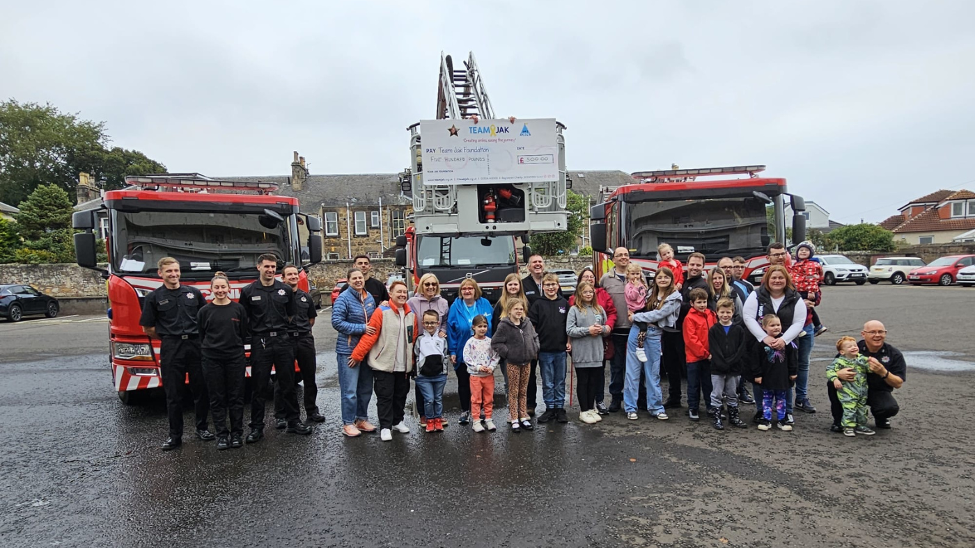 Several crew members from Kirkcaldy Fire Station with service users of Team Jak, including children and their families all standing in front of three fire engines and a cheque for £500 presented to Team Jak