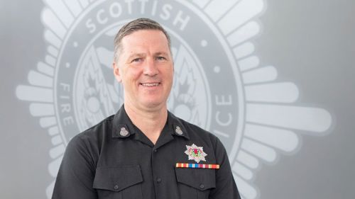 Andy Watt in black SFRS uniform standing in front of a grey SFRS crest