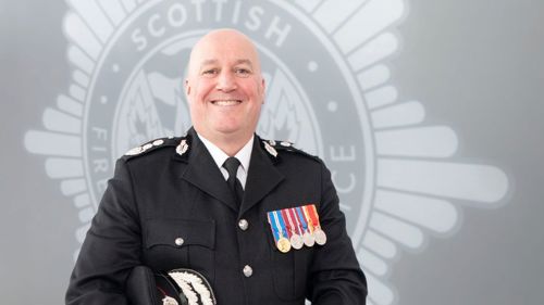 Chief Officer Ross Haggart in full uniforn standing in front of a grey SFRS crest