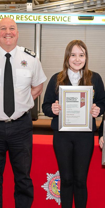 Three children receive award from top fire officer in Scotland, standing in front of fire appliance.